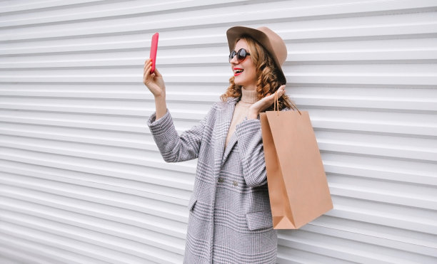 时尚快乐的笑着的女人用智能手机自拍照片，灰色外套，圆帽在城市街道上的白色墙壁背景图片下载