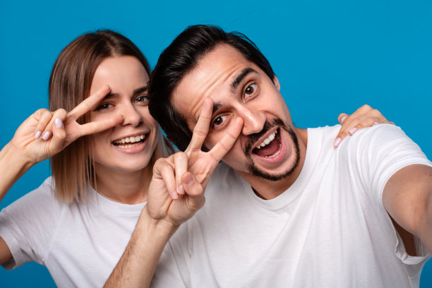 一对幸福的夫妇，一个年轻的金发女人和深色胡子的男人在白色t恤和蓝色牛仔裤上自拍孤立的蓝色背景。理想夫妻的概念。图片下载