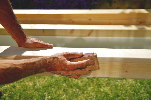 在室外工作时，男人的手和手臂用砂纸打磨天然木梁。打磨车棚用云杉木梁图片下载