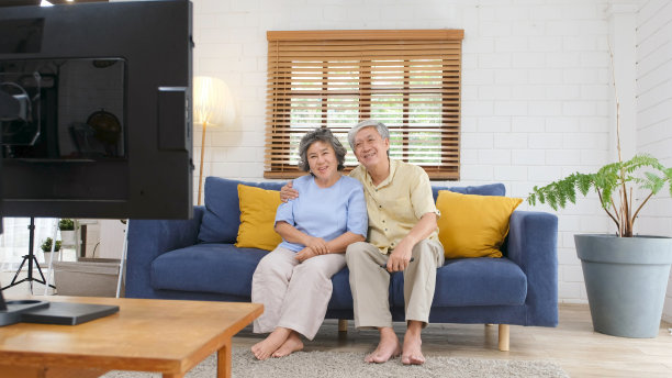 亚洲老两口一边拿着遥控器一边在家里客厅看电视，安享晚年老人的日常生活方式图片下载