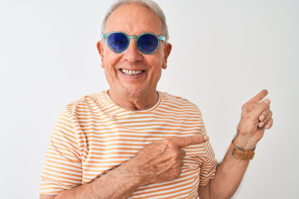 白发苍苍的老人穿着条纹t恤和太阳镜在孤立的白色背景微笑着看着摄像机，双手和手指指向一边。图片下载