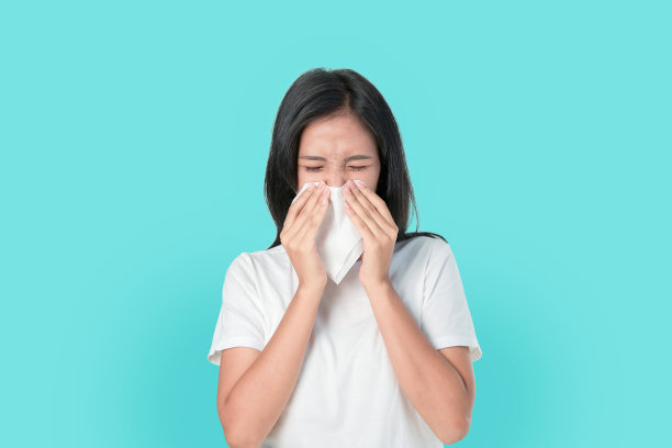 年轻的亚洲妇女使用餐巾纸的嘴和鼻子，因为过敏。在蓝色背景上。图片下载