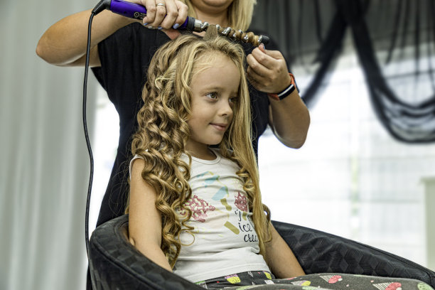 可爱的小女孩在美容院的镜子里看着自己的发型，发型师的手拿着卷发棒图片下载