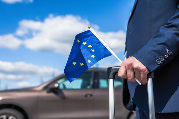 一名身穿蓝色西装、举着欧洲或欧盟旗帜的男子停在机场，他的汽车就在他的身后。商务旅行的概念图片下载