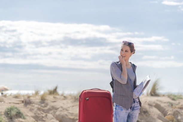 年轻的金发女郎一只手里拿着一张地图，另一只手把地图放在嘴里，一边看一边，一边拿着一只红色的手提箱，背景是天空和白云。旅行的概念。图片下载