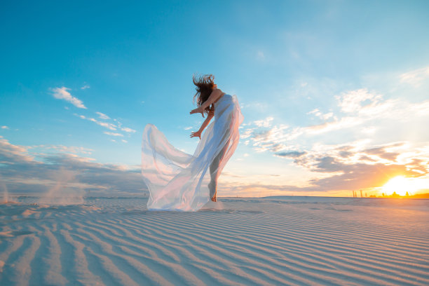 夕阳西下的沙漠中，一个穿着白裙的女孩一边跳舞一边摆姿势图片下载
