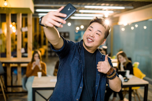 时髦的现代，开朗的中国男子在他的手机，智能手机的前摄像头拍摄自我照片，显示拇指向上的符号，站在现代阁楼的工作场所。同事会议介绍背景图片下载