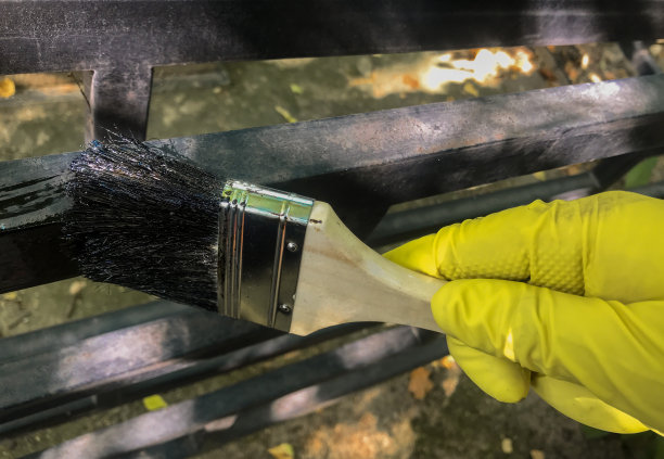 一只手在一个亮黄色橡胶手套拿着木刷和油漆黑色漆漆金属栅栏的背景自然图片下载