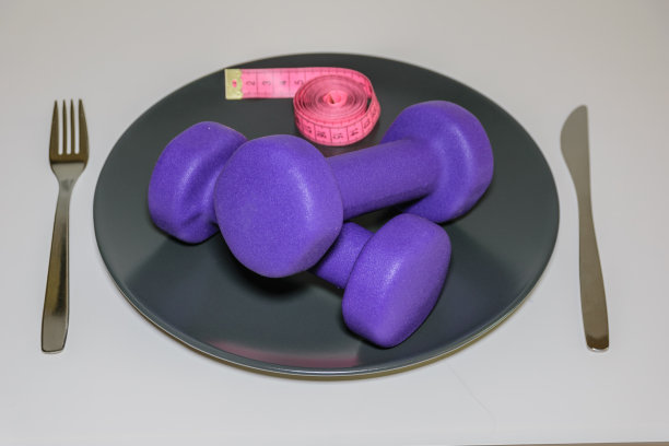 两个紫色哑铃和卷尺放在一个配有刀叉的白色盘子上，这就是健康生活方式的概念。图片下载