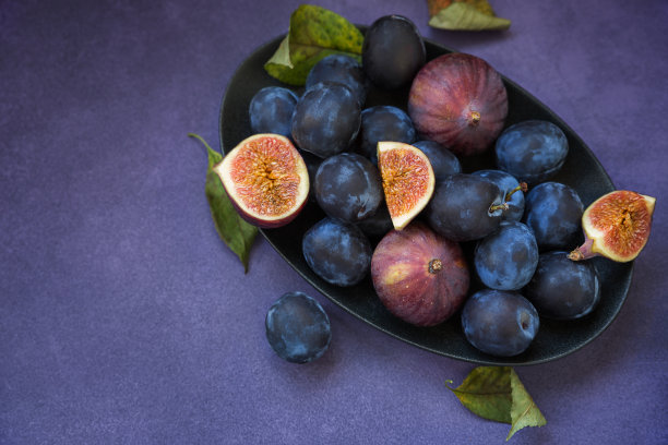 黑色的盘子和紫色的背景上放着成熟的李子和无花果。美丽的食物照片图片下载