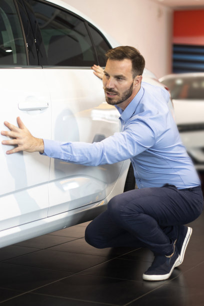一个人蹲在一辆新车旁边，而看一些特点在汽车经销商的陈列室图片下载