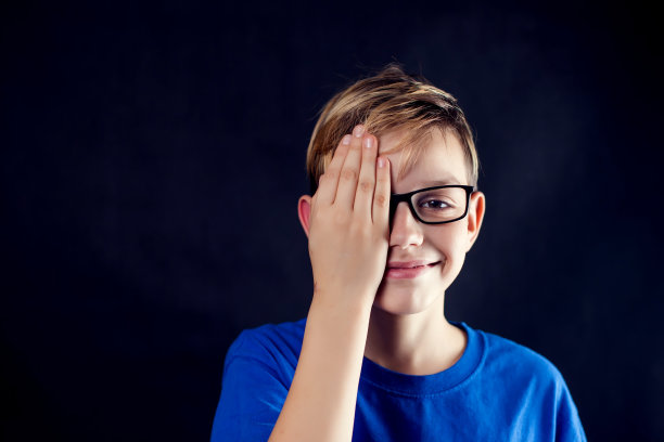 一个戴眼镜的男孩的肖像，用手掌闭上一只眼睛，在黑暗的背景前检查视力。儿童、眼科和保健概念图片下载
