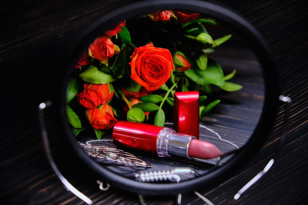 玫瑰和口红反射在镜子里。深色木质背景上的女性珠宝。一束鲜花和红色唇膏。图片下载