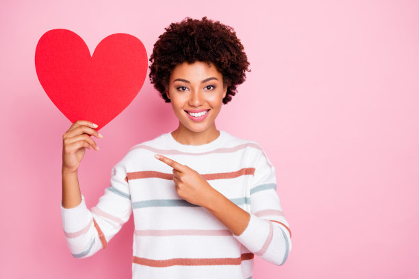 照片漂亮的深色卷曲的皮肤女士手中拿着大红色纸心建议选择创意现在穿条纹套头衫孤立的淡粉色背景图片下载