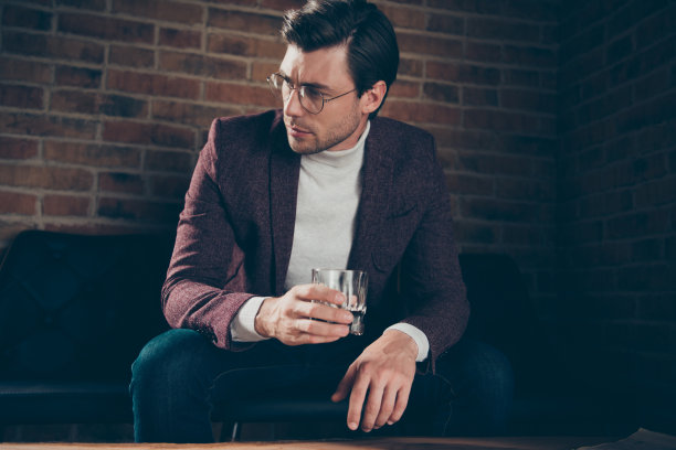 这是一张帅气的商业男人拿着酒的照片，穿着眼镜，穿着时髦的运动夹克，高领毛衣，坐在沙发上，现代的办公室图片下载