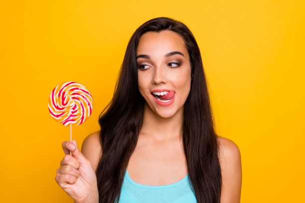 这是一张可爱可爱的小女孩的照片，她穿着蓝绿色的背心，在鲜艳的颜色背景下，看着她舔着的糖果，等待棒棒糖被吃掉图片下载