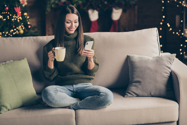 全尺寸的照片，积极开朗的女孩坐在沙发上，交叉双腿，用手机发短信，在度假屋输入圣诞信息充满了圣诞灯，室内noel装饰图片下载