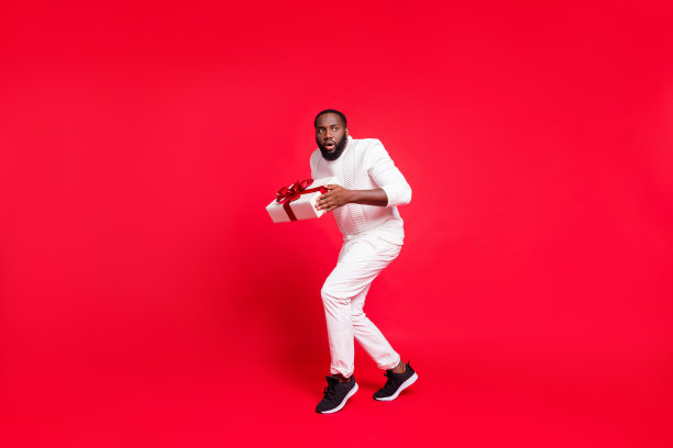 全尺寸照片有趣的黑皮肤的男人让女朋友的新年惊喜踮着脚尖携带秘密的礼物盒在圣诞树下穿毛衣裤子孤立的红色背景图片下载