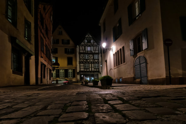 街道和旧的半木房子。傍晚时分，照亮一盏路灯。视图从下面。前景是铺路石。一辆车从后面驶过。背景。图片下载