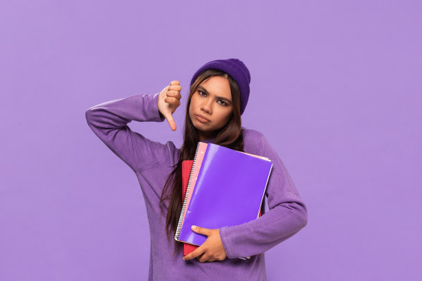 悲伤的非洲裔美国少年戴着帽子，穿着毛衣，拿着文件夹，拇指朝下，孤立地站在紫色的背景上。评价的概念。图片下载