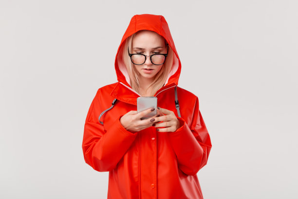 漂亮的金发女人穿着红色雨衣，带着兜帽，在白色背景下查看智能手机的通知。在一个寒冷的雨天散步图片下载