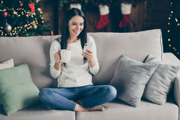 积极的女孩的完整的照片享受圣诞假期的气氛，想买梦想的礼物新年礼物，坐沙发，用智能手机，发短信，持有杯子，喝饮料，休息，在家里与noel灯图片下载