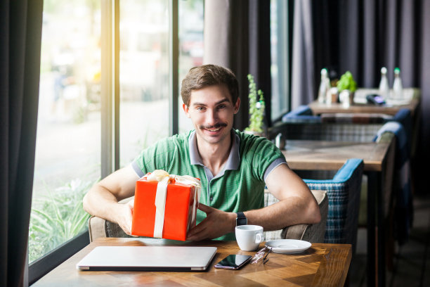 年轻快乐的商人坐在绿色t恤，拿着大红色礼品盒，看着镜头，露齿微笑。商业和自由职业的概念。图片下载