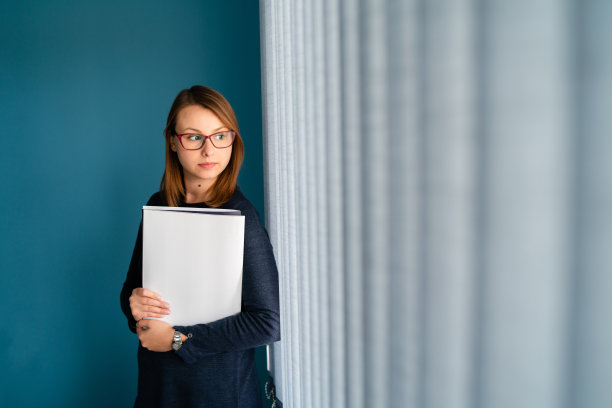 年轻女商人的肖像与眼镜秘书拿着文件文件夹工作申请前在蓝色的墙靠窗户看图片下载