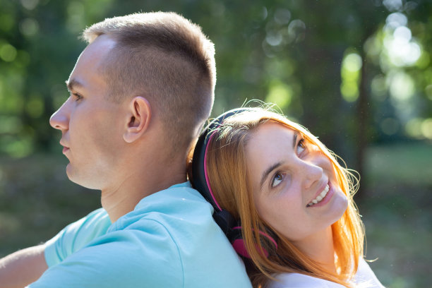 一对年轻的青少年情侣在夏天的公园里。红发女孩戴着耳机听音乐，男孩觉得很无聊。图片下载