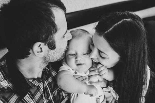 年轻微笑的家庭亲吻儿子的肖像。幸福的家庭，为人父母和人们的观念-母亲，父亲和孩子在家里。黑白照片。图片下载