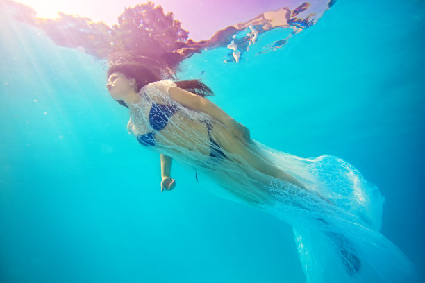 性感的年轻女孩游泳下的水在一个白色透明的披肩和蓝色泳衣，反对阳光和天空的明亮射线。数字肖像艺术。底部视图。图片下载