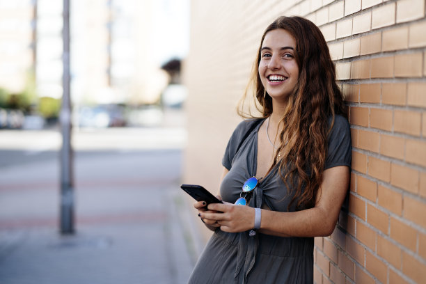 一个美丽的女人看着相机微笑和使用智能手机在街上的肖像图片下载