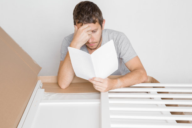 认真的年轻人阅读说明书，试图了解如何组装新家具。纸板箱里婴儿床用的白色木板。父亲为将来的孩子做准备。图片下载