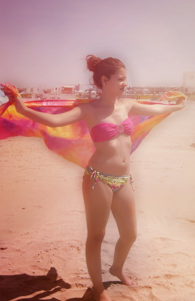 年轻女子在海滩上穿着比基尼伸展织物在风在她的背后，双臂图片下载