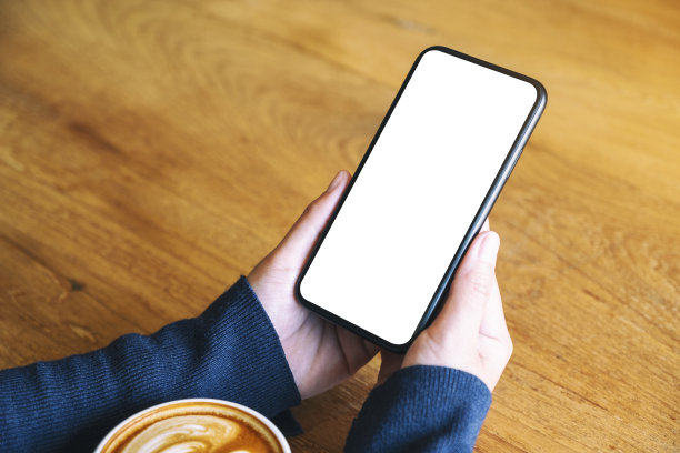 一个女人拿着白色的手机和空白屏幕，咖啡杯放在木桌上图片下载