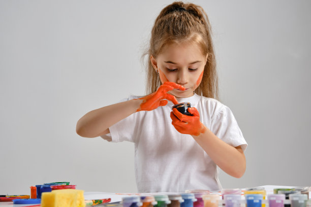 一个穿着白色t恤的小女孩坐在桌子旁边，桌上画着什么人和五颜六色的颜料，画着她的手。孤立在白色的。媒介特写图片下载