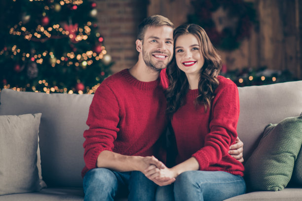 新年快乐。在装饰的花环灯房间里，情绪的夫妇在室内穿着红色套头衫坐在舒适的沙发上，迎接圣诞节的照片图片下载