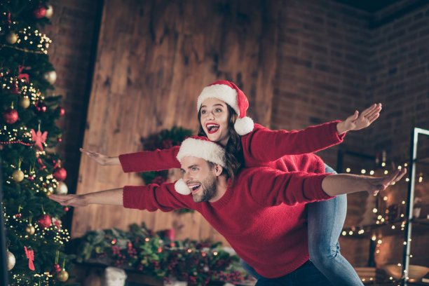 有趣的夫妇在装饰的花环灯房间里度过圣诞前夜的照片，男人背着女士假装飞行穿着红色套头衫和圣诞老人的帽子在室内图片下载