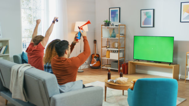 三个体育迷坐在客厅的沙发上看绿色的彩色键屏幕电视重要的比赛，使用喇叭，有乐趣和为他们的球队欢呼。图片下载