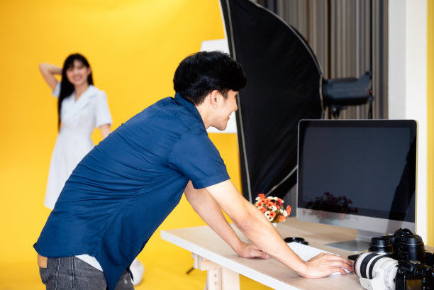 在幕后，摄影师正在检查照片，美丽的年轻女模特微笑着在工作室的背景和灯光摆姿势。有选择性的重点图片下载