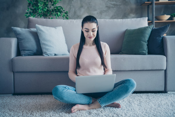 照片上的漂亮女士拿着笔记本在家里工作，靠在沙发上，坐在地板上舒适的蓬松的地毯上，穿着套衫和牛仔裤在公寓图片下载