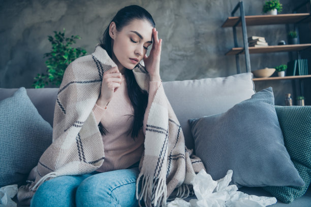 照片疲惫的女士感觉不好，触摸受伤的头部，覆盖着毯子，穿着套衫和牛仔裤坐在沙发周围许多纸餐巾图片下载