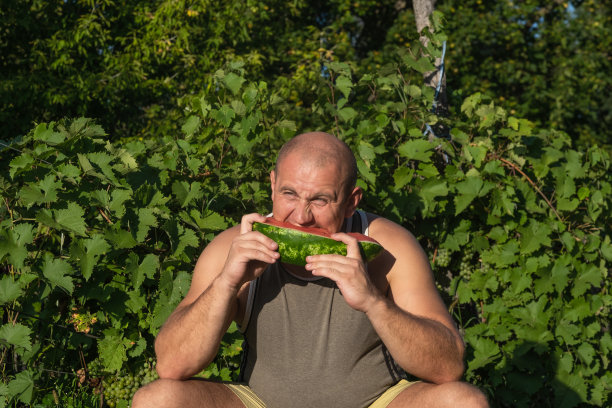 残忍的男人享受着甜甜的红西瓜。35岁男模坐在自然绿色的背景下吃新鲜水果。快乐的吃人。图片下载