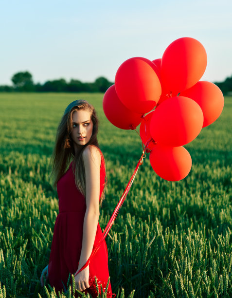 年轻漂亮的女人穿着红色的裙子摆姿势在绿色的田野与红色的气球图片下载