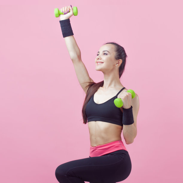 一个年轻的运动女性，带着微笑，穿着黑色的运动上衣，在粉红色的背景上做有氧运动，绿色的光哑铃。图片下载