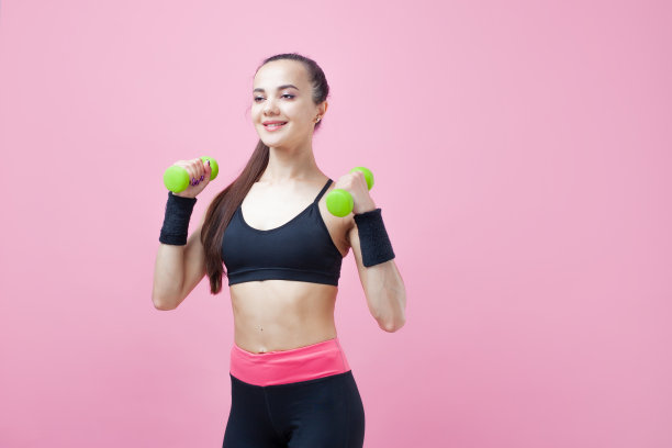 一个年轻的运动女性，带着微笑，穿着黑色的运动上衣，在粉红色的背景上做有氧运动，绿色的光哑铃。图片下载
