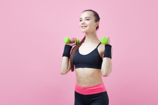 一个年轻的运动女性，带着微笑，穿着黑色的运动上衣，在粉红色的背景上做有氧运动，绿色的光哑铃。图片下载