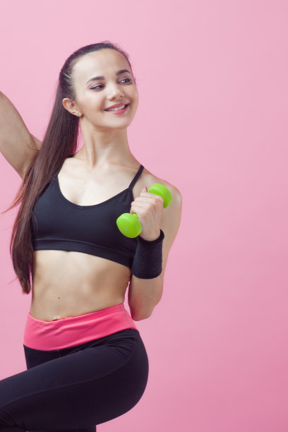 一个年轻的运动女性，带着微笑，穿着黑色的运动上衣，在粉红色的背景上做有氧运动，绿色的光哑铃。图片下载