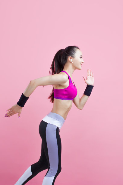 一个年轻迷人的女孩微笑着跑着，戴着无线耳机，穿着明亮的运动上衣，粉红色的背景，有氧运动。图片下载