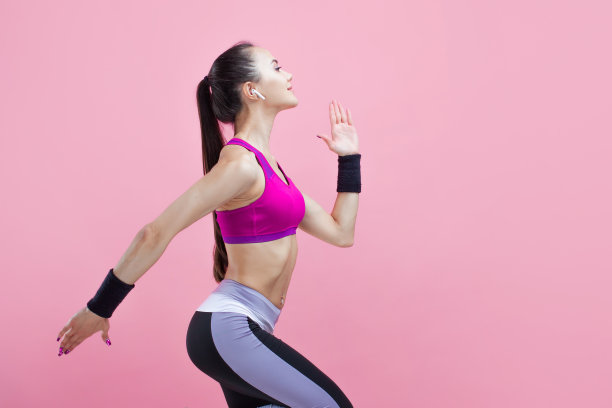 一个年轻迷人的女孩微笑着跑着，戴着无线耳机，穿着明亮的运动上衣，粉红色的背景，有氧运动。图片下载